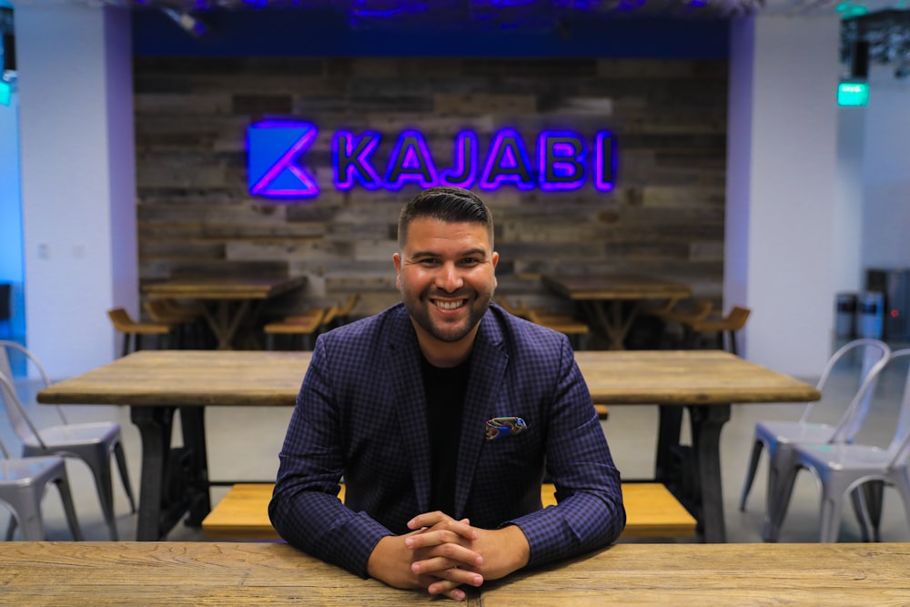 man smiling and sitting beside table in front of Kajabi neon signage turned on