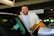 A man with a beard and wearing a light-colored shirt stands near the open door of a car in a parking garage. He is smiling broadly, creating a cheerful atmosphere. The garage ceiling and signage indicating directions to a terminal are visible, creating a busy environment.