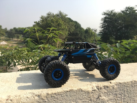A remote controlled toy truck is placed on a concrete surface, against a backdrop of lush green foliage. The vehicle has large, rugged tires and a sleek black and blue body, designed in a style reminiscent of a rock crawler. Sunlight casts a shadow from the toy onto the concrete, highlighting the details of its structure. In the distance, there are more trees and a clear sky.