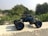 A remote controlled toy truck is placed on a concrete surface, against a backdrop of lush green foliage. The vehicle has large, rugged tires and a sleek black and blue body, designed in a style reminiscent of a rock crawler. Sunlight casts a shadow from the toy onto the concrete, highlighting the details of its structure. In the distance, there are more trees and a clear sky.