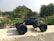 A remote controlled toy truck is placed on a concrete surface, against a backdrop of lush green foliage. The vehicle has large, rugged tires and a sleek black and blue body, designed in a style reminiscent of a rock crawler. Sunlight casts a shadow from the toy onto the concrete, highlighting the details of its structure. In the distance, there are more trees and a clear sky.