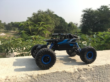 A remote controlled toy truck is placed on a concrete surface, against a backdrop of lush green foliage. The vehicle has large, rugged tires and a sleek black and blue body, designed in a style reminiscent of a rock crawler. Sunlight casts a shadow from the toy onto the concrete, highlighting the details of its structure. In the distance, there are more trees and a clear sky.