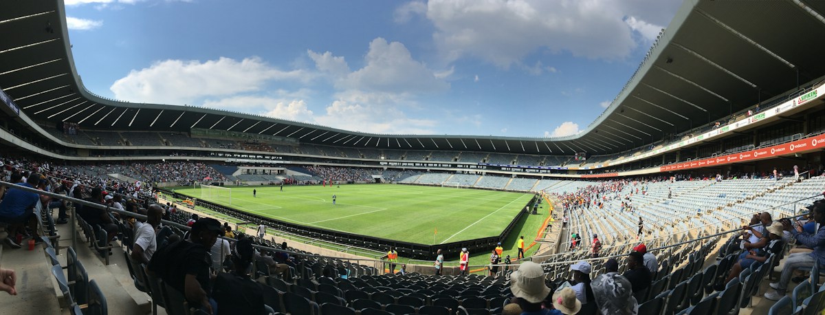 Des spectateurs suivent un match de football depuis les tribunes d’un grand stade, sous un ciel bleu et nuageux