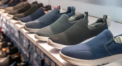 A row of slip-on shoes is neatly arranged on a white shelf, showcasing various designs and colors including blue, black, and dark grey. The shoes have a casual style and appear to be made of breathable fabric with textured patterns. The display creates a sense of order and organization within a retail setting.
