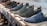 A row of slip-on shoes is neatly arranged on a white shelf, showcasing various designs and colors including blue, black, and dark grey. The shoes have a casual style and appear to be made of breathable fabric with textured patterns. The display creates a sense of order and organization within a retail setting.