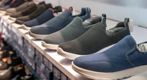A row of slip-on shoes is neatly arranged on a white shelf, showcasing various designs and colors including blue, black, and dark grey. The shoes have a casual style and appear to be made of breathable fabric with textured patterns. The display creates a sense of order and organization within a retail setting.
