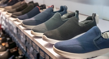 A row of slip-on shoes is neatly arranged on a white shelf, showcasing various designs and colors including blue, black, and dark grey. The shoes have a casual style and appear to be made of breathable fabric with textured patterns. The display creates a sense of order and organization within a retail setting.