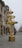 A tall, decorative installation resembling a Christmas tree stands on a city street. It is adorned with numerous small, warm white lights wrapped around its structure, which is tiered with garland-like decorations. The installation is placed on a large cylindrical base. In the background, there is a historic building with ornate architectural details. The street appears empty, with a few people walking in the distance.