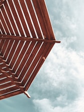 A perspective of a wooden pergola with slatted beams set against a partly cloudy sky.