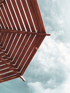A perspective of a wooden pergola with slatted beams set against a partly cloudy sky.