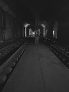 A dimly lit underground tunnel with two sets of railway tracks running parallel. The tunnel walls are concrete, and there are lights along the right side, casting shadows and creating a sense of depth. The end of the tunnel is faintly visible in the distance.