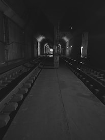 A dimly lit underground tunnel with two sets of railway tracks running parallel. The tunnel walls are concrete, and there are lights along the right side, casting shadows and creating a sense of depth. The end of the tunnel is faintly visible in the distance.