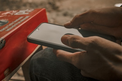 A person holds a smartphone with both hands, focusing intently on the screen. To the side, there is a red book with text partially visible. The overall setting is informal and intimate, suggesting a moment of concentration or relaxation.