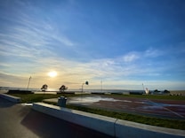 A serene coastal scene with a basketball court, bordered by lush green grass. The sun is setting over the ocean, casting a warm glow on the surroundings, while the sky is painted in hues of blue and orange. A few trees and lampposts are in the area, enhancing the peaceful ambiance.