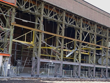 A large industrial building with a steel framework, featuring rust and wear. The structure includes multiple levels supported by metal beams, with exposed wiring and various construction materials scattered on the site. Safety barriers and fencing indicate an area under construction or repair.