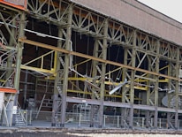 A large industrial building with a steel framework, featuring rust and wear. The structure includes multiple levels supported by metal beams, with exposed wiring and various construction materials scattered on the site. Safety barriers and fencing indicate an area under construction or repair.