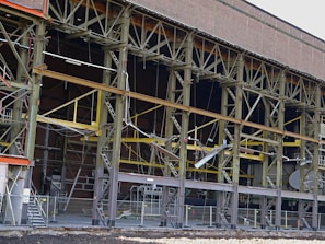 A large industrial building with a steel framework, featuring rust and wear. The structure includes multiple levels supported by metal beams, with exposed wiring and various construction materials scattered on the site. Safety barriers and fencing indicate an area under construction or repair.