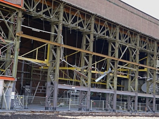 A large industrial building with a steel framework, featuring rust and wear. The structure includes multiple levels supported by metal beams, with exposed wiring and various construction materials scattered on the site. Safety barriers and fencing indicate an area under construction or repair.