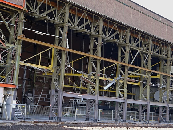 A large industrial building with a steel framework, featuring rust and wear. The structure includes multiple levels supported by metal beams, with exposed wiring and various construction materials scattered on the site. Safety barriers and fencing indicate an area under construction or repair.