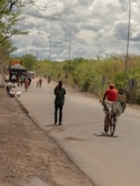 A rural road scene features several people walking and cycling along a paved road bordered by vegetation. A person wearing a red hat rides a bicycle carrying boxes. A group of people and some vehicles are seen in the background, while the sky above is filled with thick, cloudy formations.