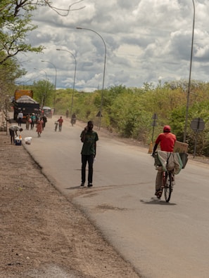 A rural road scene features several people walking and cycling along a paved road bordered by vegetation. A person wearing a red hat rides a bicycle carrying boxes. A group of people and some vehicles are seen in the background, while the sky above is filled with thick, cloudy formations.