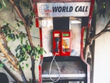 An old, worn-out telephone booth with a red payphone placed against a backdrop of a wall and some leafy tree branches. The sign above the payphone reads 'World Call' with a globe logo. The booth has visible signs of wear, including peeling paint and scratches.