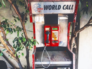 An old, worn-out telephone booth with a red payphone placed against a backdrop of a wall and some leafy tree branches. The sign above the payphone reads 'World Call' with a globe logo. The booth has visible signs of wear, including peeling paint and scratches.