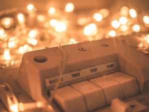 A close-up of an old audio device with buttons labeled 'Record,' 'Trick,' 'Stop,' and 'Start.' The device is surrounded by warm, glowing fairy lights that create a soft bokeh effect in the background.