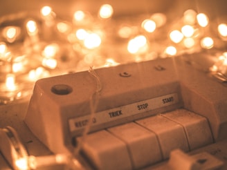 A close-up of an old audio device with buttons labeled 'Record,' 'Trick,' 'Stop,' and 'Start.' The device is surrounded by warm, glowing fairy lights that create a soft bokeh effect in the background.