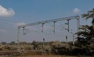 Overhead railway electrical lines cross a rural landscape, with sturdy metal poles supporting a network of wires. The area is surrounded by dense vegetation and the sky is clear with few clouds.