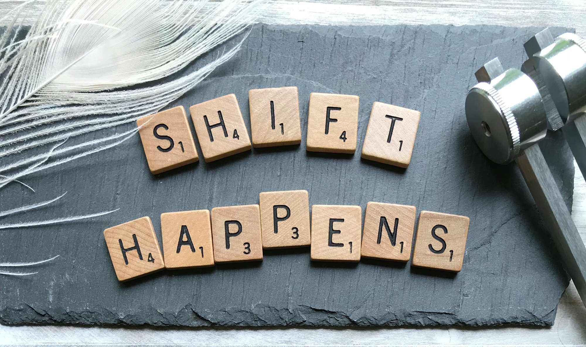letter tiles spelling out "shift happens" on a gray surface with a feather to the left and metal to the right