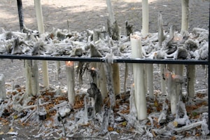 A scene of melted and partially melted candles on a black metal stand. The wax appears to have dripped extensively, creating intricate patterns and structures. Some candles are still burning, with small flames visible. The surrounding ground is covered in wax drippings and fallen leaves, adding texture to the scene. The background features a stone pavement surface.