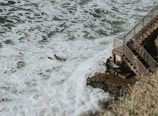 A surfer is paddling on a surfboard through white, foamy waves near a rocky coastline. Another surfer with a board is standing on a rock next to a metal railing staircase leading down to the water. The scene conveys a sense of adventure and connection to nature.