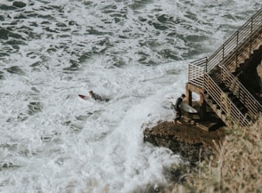A surfer is paddling on a surfboard through white, foamy waves near a rocky coastline. Another surfer with a board is standing on a rock next to a metal railing staircase leading down to the water. The scene conveys a sense of adventure and connection to nature.