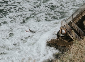 A surfer is paddling on a surfboard through white, foamy waves near a rocky coastline. Another surfer with a board is standing on a rock next to a metal railing staircase leading down to the water. The scene conveys a sense of adventure and connection to nature.