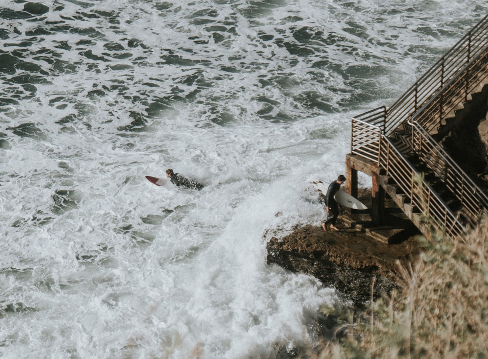 ocean wave crashing to rocky shore with man carrying surfboard beside stairs