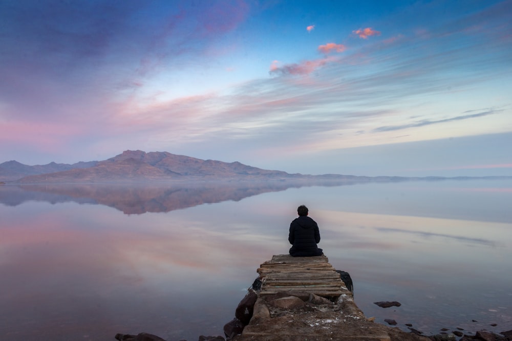 man sitting on cliff looking at body of water during golden hour