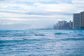 Surfers navigate through small to medium waves in the ocean while high-rise buildings line the shore in the background. The sky is overcast with a blend of white and gray clouds.