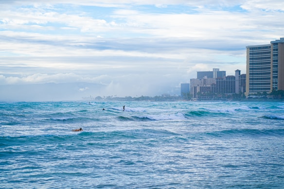 Surfers navigate through small to medium waves in the ocean while high-rise buildings line the shore in the background. The sky is overcast with a blend of white and gray clouds.