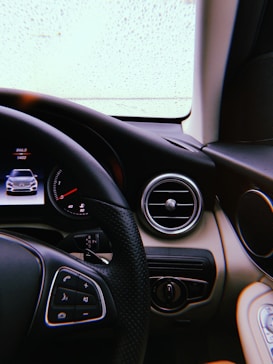 The interior view of a car focusing on the steering wheel and dashboard. The dashboard features a digital display showing a car image and various controls, while the steering wheel has several buttons for managing functions. Raindrops can be seen on the windshield, indicating wet weather outside.