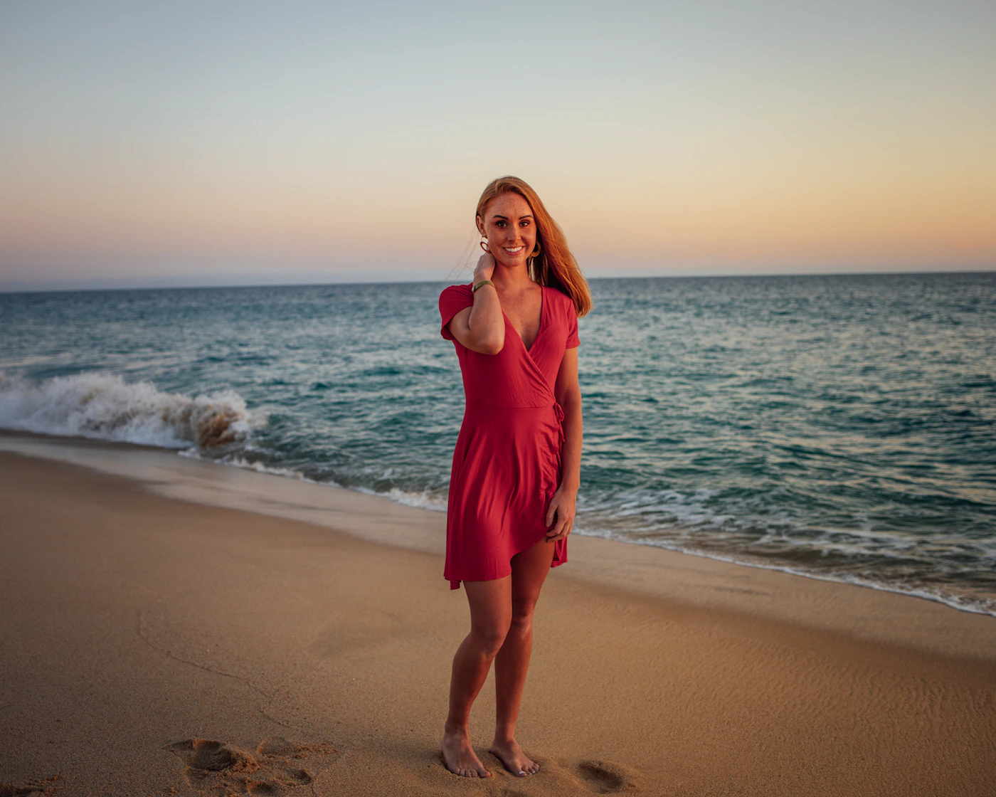 A woman in a red dress standing on the Cabo San Lucas beach at golden hour, the sky fading from pale blue to warm orange above the calm ocean, warm light wrapping across the sand