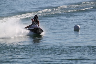 A person riding a jet ski at high speed creates a large splash on a body of water. The water is calm with gentle waves, and there is a buoy floating nearby.