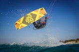A person is performing a high-flying kitesurfing trick over the ocean, captured mid-air against a clear blue sky. The kitesurfer is wearing a wetsuit and holding onto the kite's handle, with the yellow and black board prominently visible. Water splashes around the surfer, adding to the dynamic action of the scene.