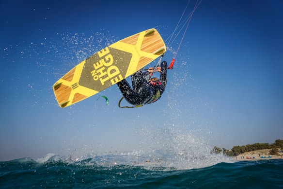 A person is performing a high-flying kitesurfing trick over the ocean, captured mid-air against a clear blue sky. The kitesurfer is wearing a wetsuit and holding onto the kite's handle, with the yellow and black board prominently visible. Water splashes around the surfer, adding to the dynamic action of the scene.