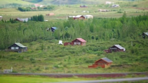 A rural landscape features scattered wooden cabins nestled among lush greenery, with a backdrop of gently rolling hills. A white vehicle is parked near one of the cabins, suggesting habitation. The serene environment is characterized by dense forest areas and open grass fields under a clear sky.