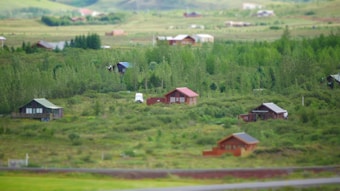 A rural landscape features scattered wooden cabins nestled among lush greenery, with a backdrop of gently rolling hills. A white vehicle is parked near one of the cabins, suggesting habitation. The serene environment is characterized by dense forest areas and open grass fields under a clear sky.