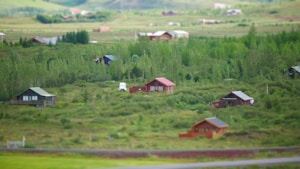 A rural landscape features scattered wooden cabins nestled among lush greenery, with a backdrop of gently rolling hills. A white vehicle is parked near one of the cabins, suggesting habitation. The serene environment is characterized by dense forest areas and open grass fields under a clear sky.