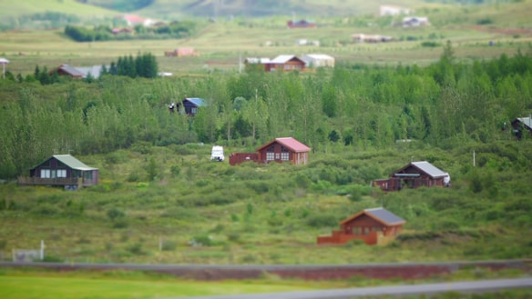A rural landscape features scattered wooden cabins nestled among lush greenery, with a backdrop of gently rolling hills. A white vehicle is parked near one of the cabins, suggesting habitation. The serene environment is characterized by dense forest areas and open grass fields under a clear sky.