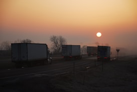 Several trucks are driving along a highway during an early morning or late evening with a hazy, misty atmosphere. The sun is low on the horizon, casting a warm, orange glow over the scene and reflecting off the road.