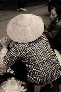 A person wearing a traditional conical hat is seated on a stool, facing away from the camera. The individual is dressed in a checkered shirt and is engaged in an activity, possibly involving plastic bags and items on the ground. Another person is partially visible in the background, sitting on the ground, attentively involved in the same activity.